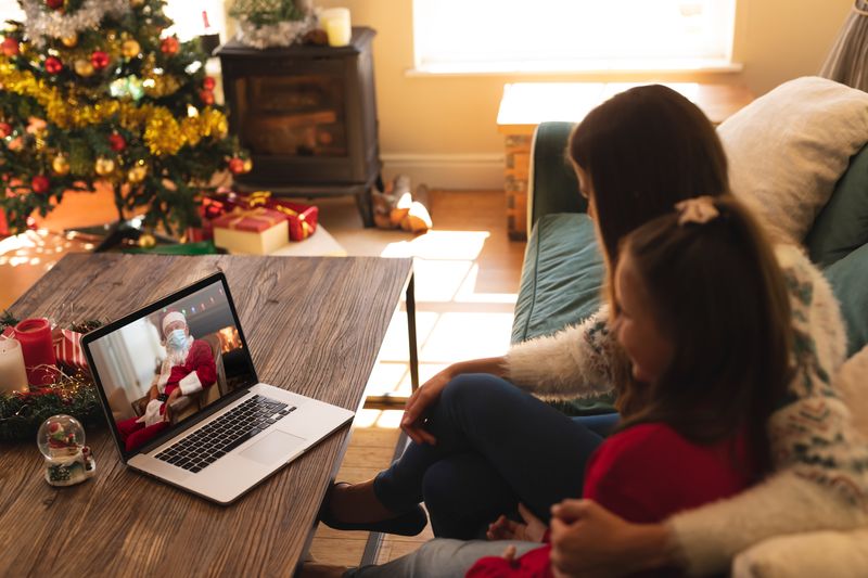 Woman and daughter sitting on the couch having a videocall with santa claus wearing face mask on lap