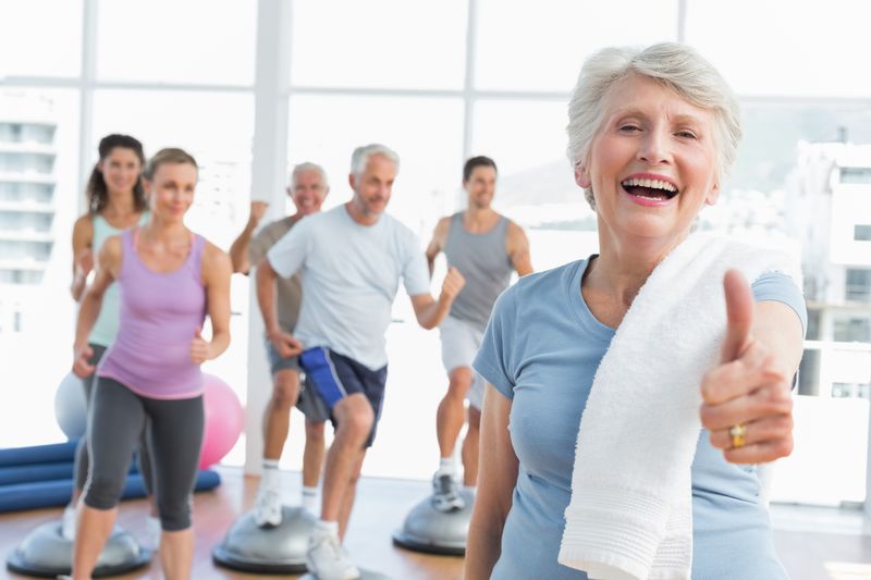 Cheerful senior woman gesturing thumbs up with people exercising in the background at fitness studio