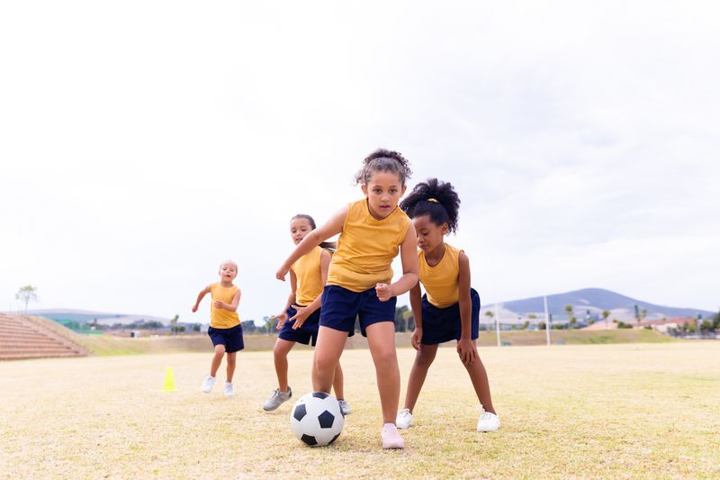 Full length of multiracial elementary schoolgirls in sports uniform playing soccer against sky. unaltered, childhood, education, sports training and sports activity concept.