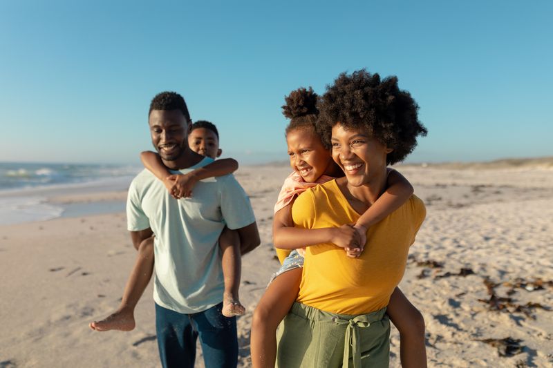 Happy african american parents giving piggyback to children while walking at beach on sunny day. unaltered, family, lifestyle, togetherness, enjoyment and holiday concept.