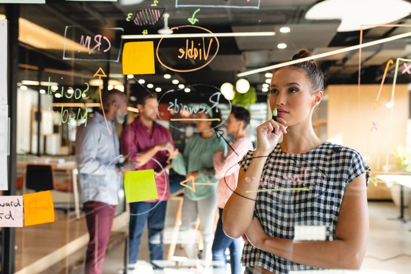 Biracial businesswoman analyzing charts on glass wall while colleagues discussing in background. Unaltered, creative business, ideas, meeting, diversity, strategy, business plan.