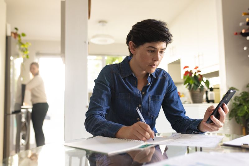 Caucasian mid adult lesbian woman with short hair using mobile phone and writing in book on table. Unaltered, technology, paperwork, business, technology, freelancer and home office concept.