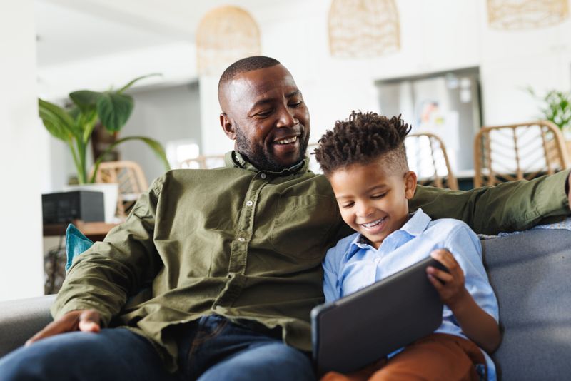 Happy african american father and son sitting on sofa, using tablet in living room. Spending quality time at home, domestic life and family concept.