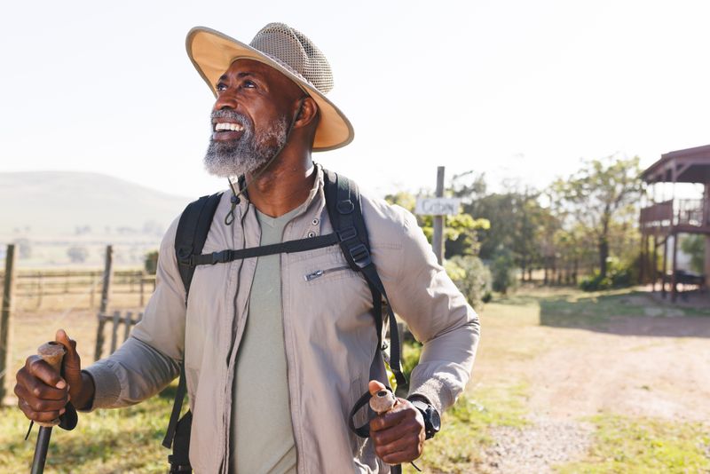 Smiling african american senior man with hiking poles standing on land against clear sky. Unaltered, copy space, hat, retirement, vacation, solitude, holiday, beard, adventure and active lifestyle.