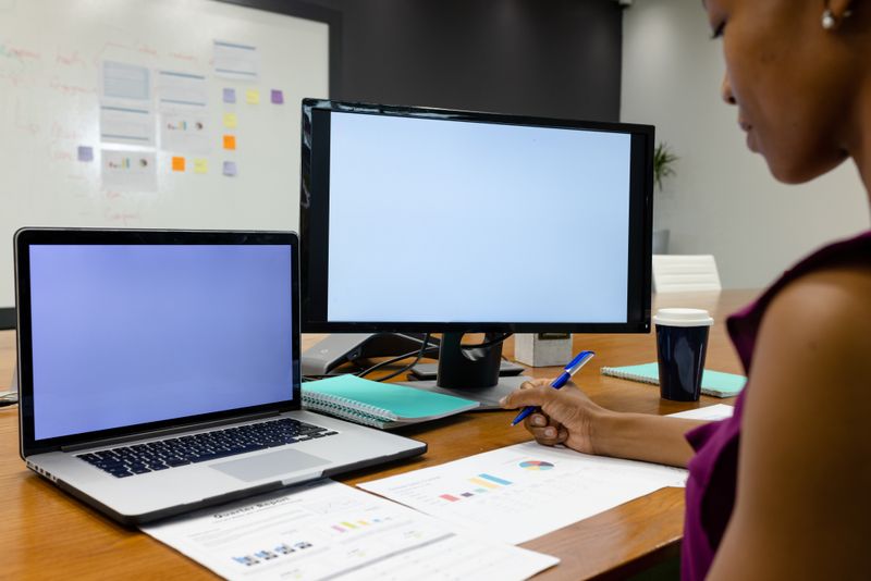 Midsection of african american businesswoman analyzing reports by laptop and computer on desk. Copy space, unaltered, architecture, technology, sales, progress, business, technology, office concept.