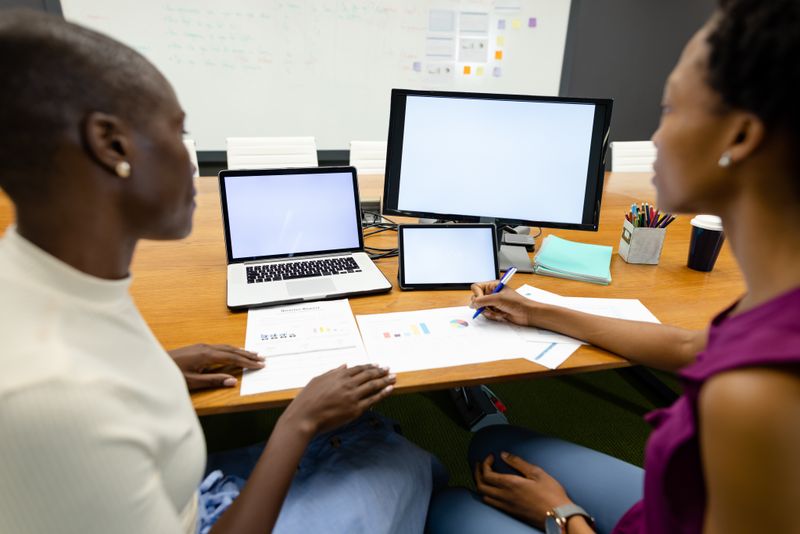 African american females colleagues analyzing sales reports by laptop and computer on desk. Copy space, unaltered, architecture, technology, sales, progress, business, technology, office concept.