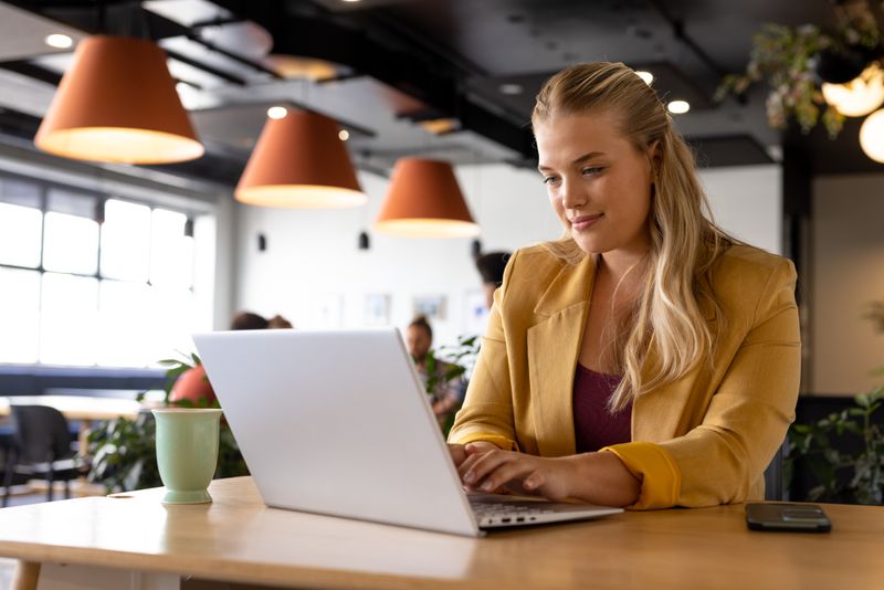 Happy plus size caucasian casual businesswoman using laptop at desk. Casual office, communication, business and work, unaltered.