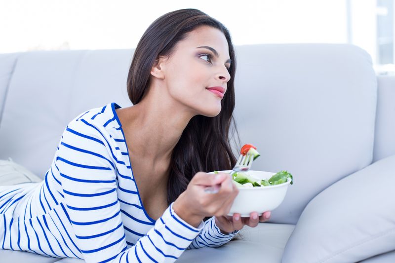 Smiling beautiful brunette relaxing on the couch and eating salad