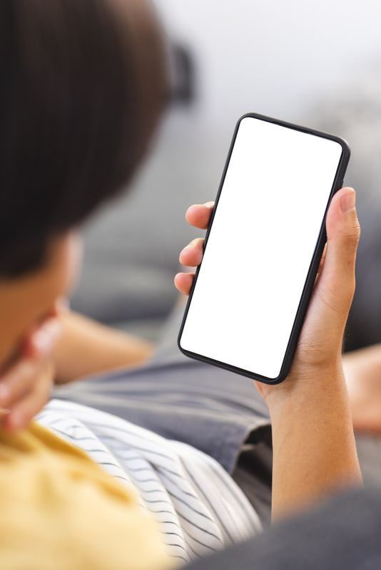 Asian college student holding smartphone with blank screen. Wearing yellow shirt, relaxing on sofa, looking at device, unaltered