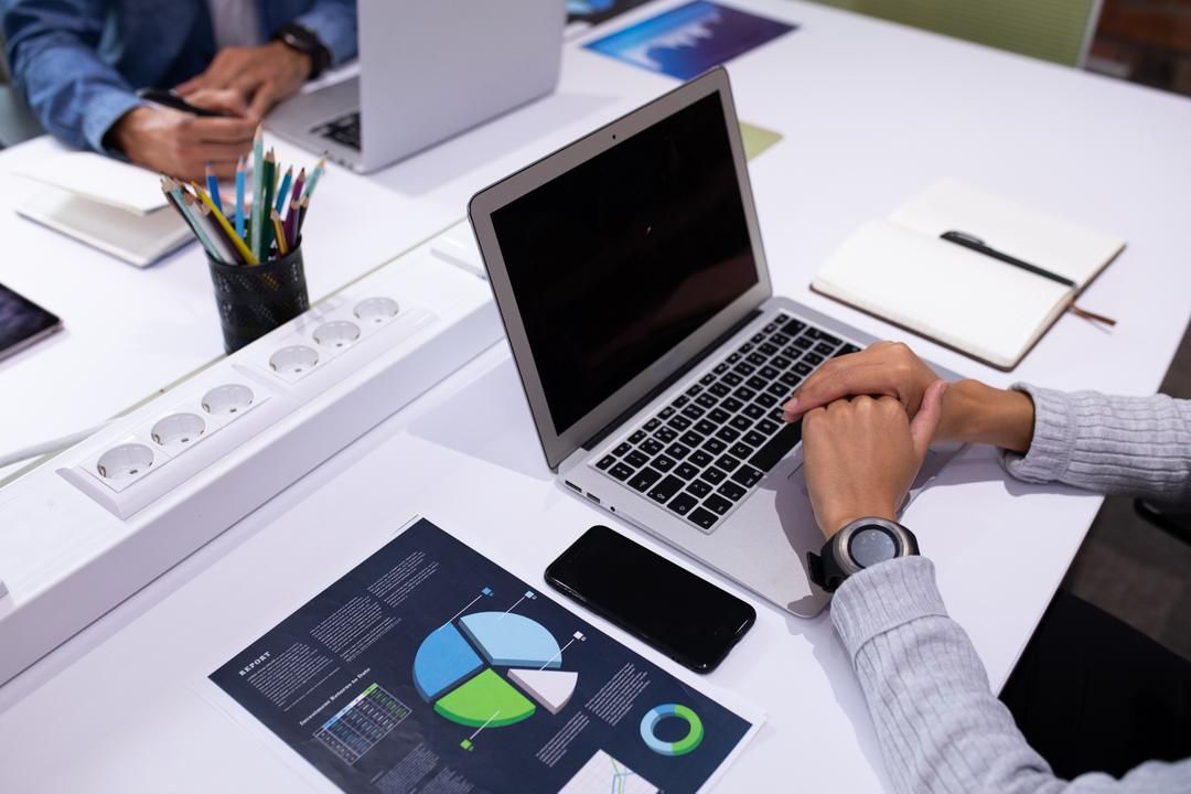 Over the shoulder image of a person sitting in front of a laptop with a phone, poster and notebook beside it - Why you should keep your posts on brand - Image