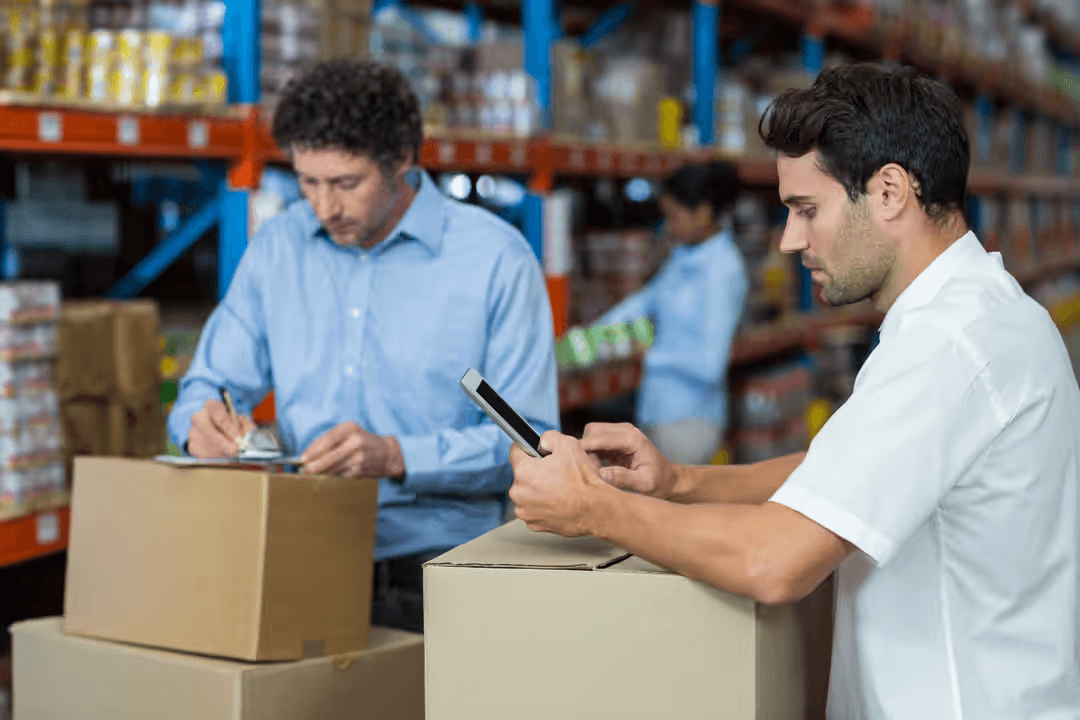 Two workers leaning on boxes in a warehouse with one looking at tablet and one writing on clipboard - Retail marketing strategy tips and tricks to drive sales and save money - Image
