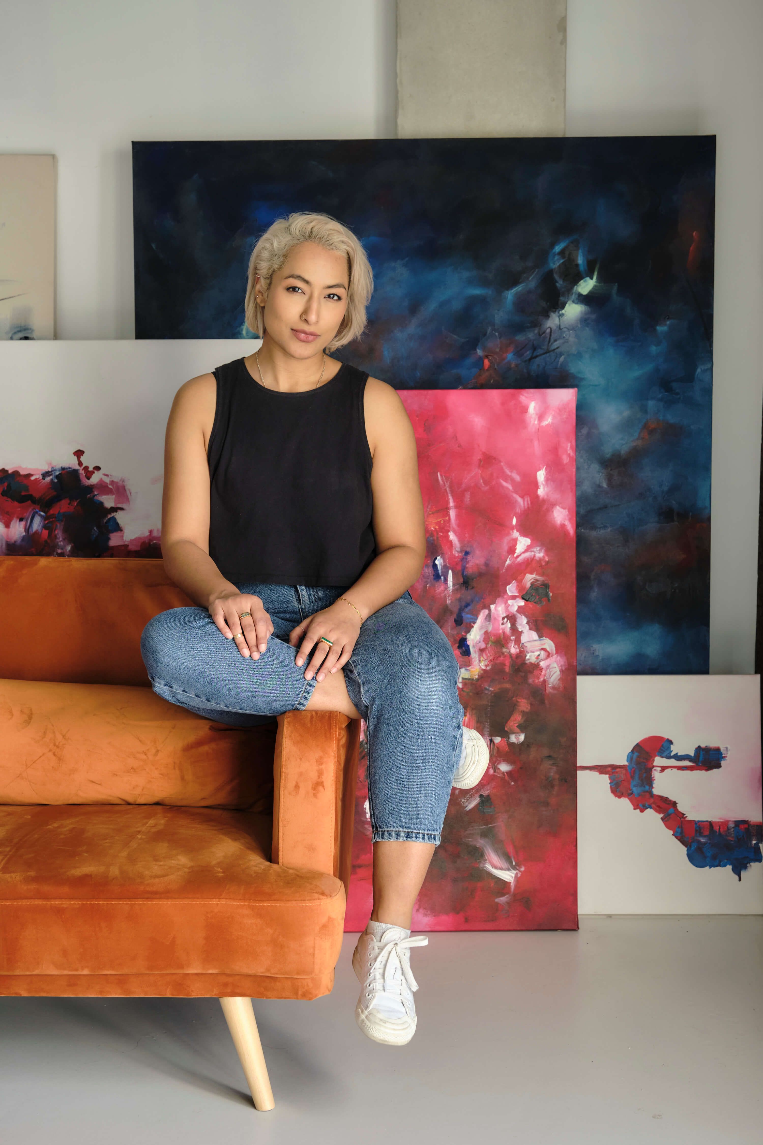 A woman artist sits on a sofa in front of her paintings - What does a maximalist design style give you that minimalism doesn't - Image