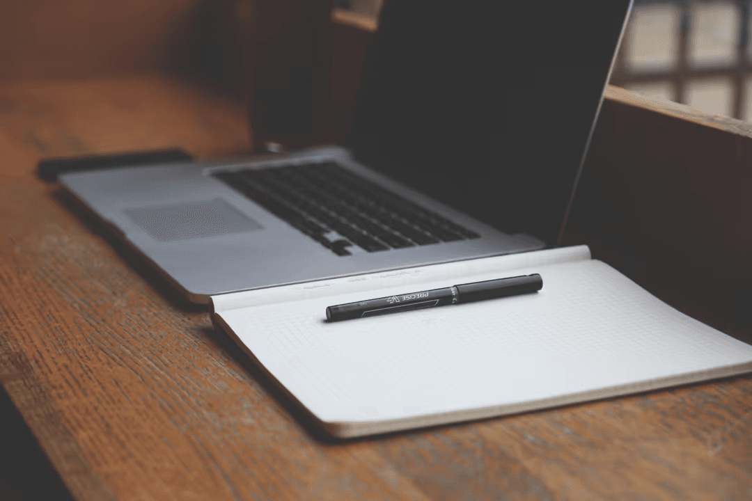 Image of a computer, notebook and pen on a wooden desk - How to make a beautiful digital vision board with free online tools in 5 steps - Image