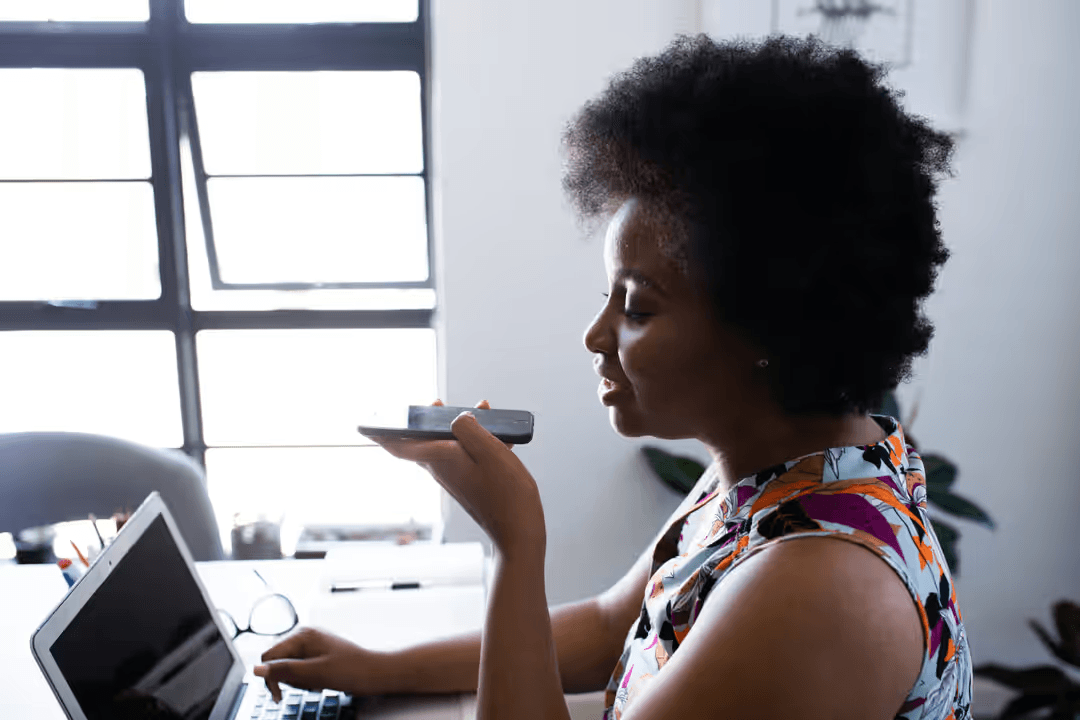 Side of a woman speaking to her phone, with her laptop in front of her on a desk - Expert online marketing tips to improve your digital strategy and make more money - Image