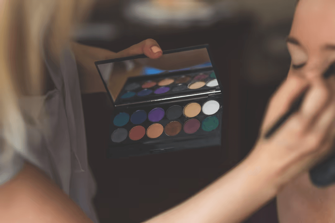 Image of a woman getting her makeup done, with applier holding an eyeshadow palette thats in focus - 35 creative YouTube video ideas when you're out of inspiration - Image