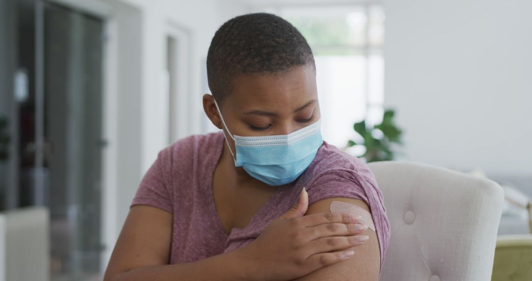 Woman with mask examining bandage on arm indoors - Free Images, Stock Photos and Pictures on Pikwizard.com