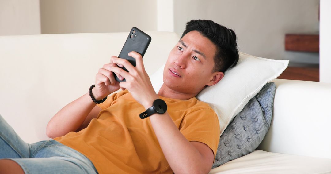 Asian Man Relaxing on Sofa Engaged with Smartphone and Wearable Tech