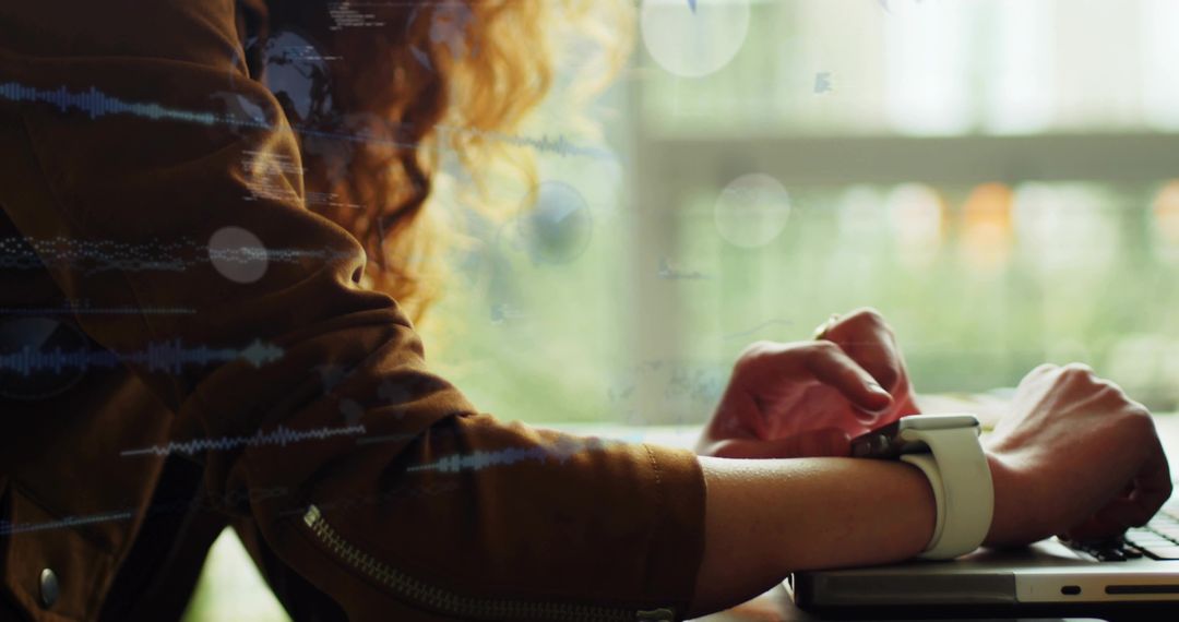 Curly-haired Woman Wearing Smartwatch Typing on Laptop Near Window with Green Bokeh
