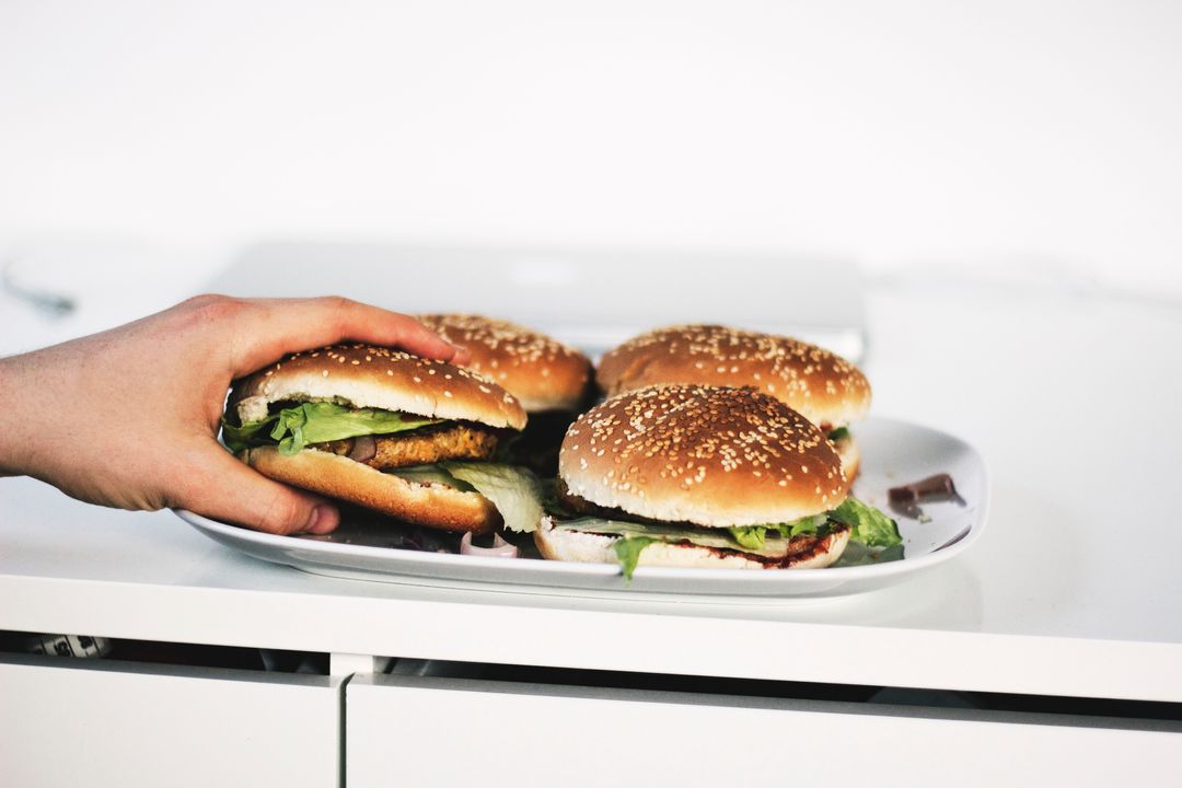 Hand Reaching for Sesame Seed Burger on Plate with Multiple Lettuce Burgers on White Table