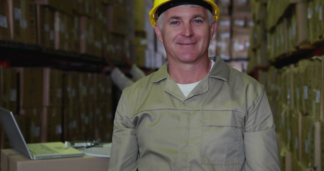 Smiling warehouse supervisor wearing yellow hard hat standing in pallet racking aisle with laptop