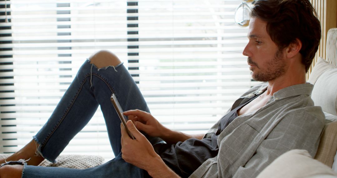 Man Relaxing at Home Reading on Digital Tablet by Window in Casual Outfit - Free Images, Stock Photos and Pictures on Pikwizard.com