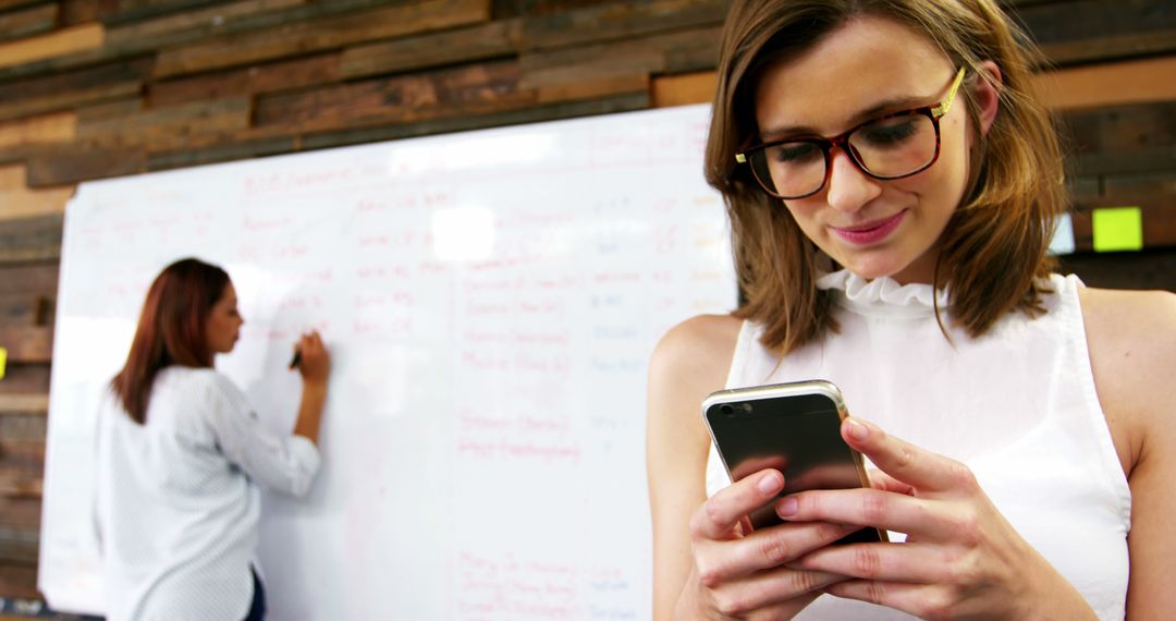 Young Professional Woman in Glasses Using Smartphone While Colleague Writes on Whiteboard - Free Images, Stock Photos and Pictures on Pikwizard.com