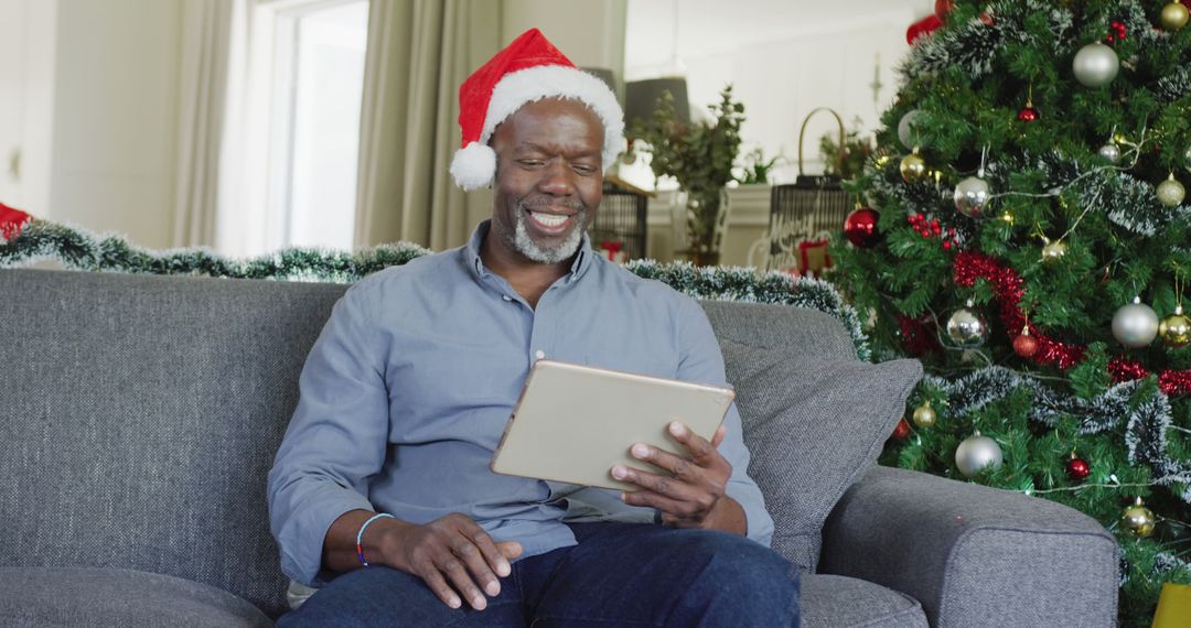 Happy Man in Santa Hat Using Tablet Next to Christmas Tree - Free Images, Stock Photos and Pictures on Pikwizard.com