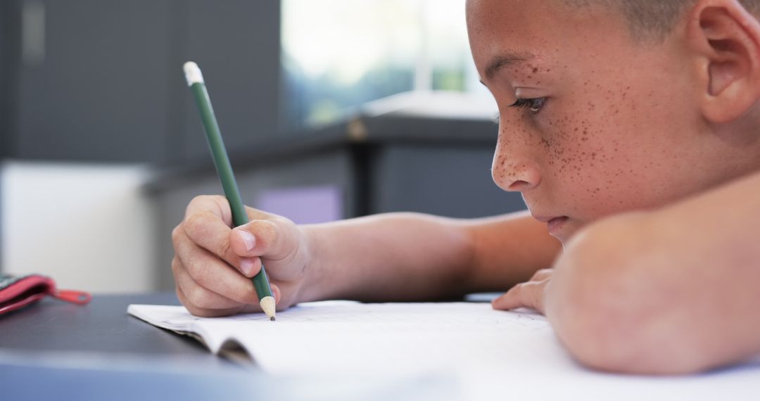 Focused Boy with Freckles Writing in Classroom Notebook