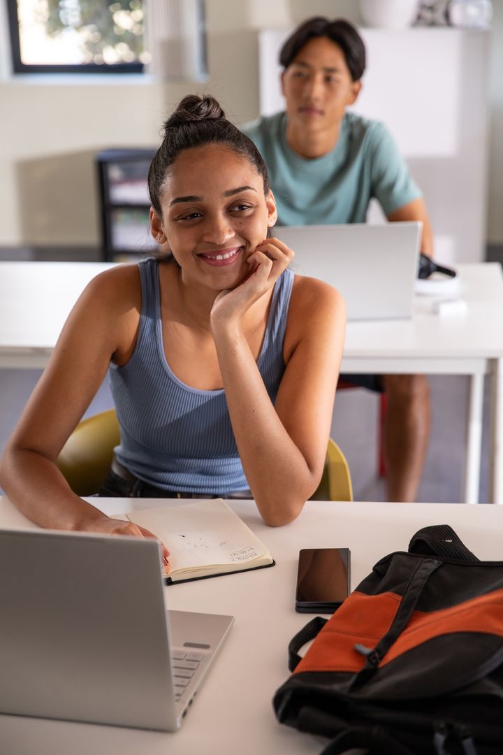Smiling teenage student studying with laptop and notebook while diverse classmates collaborate