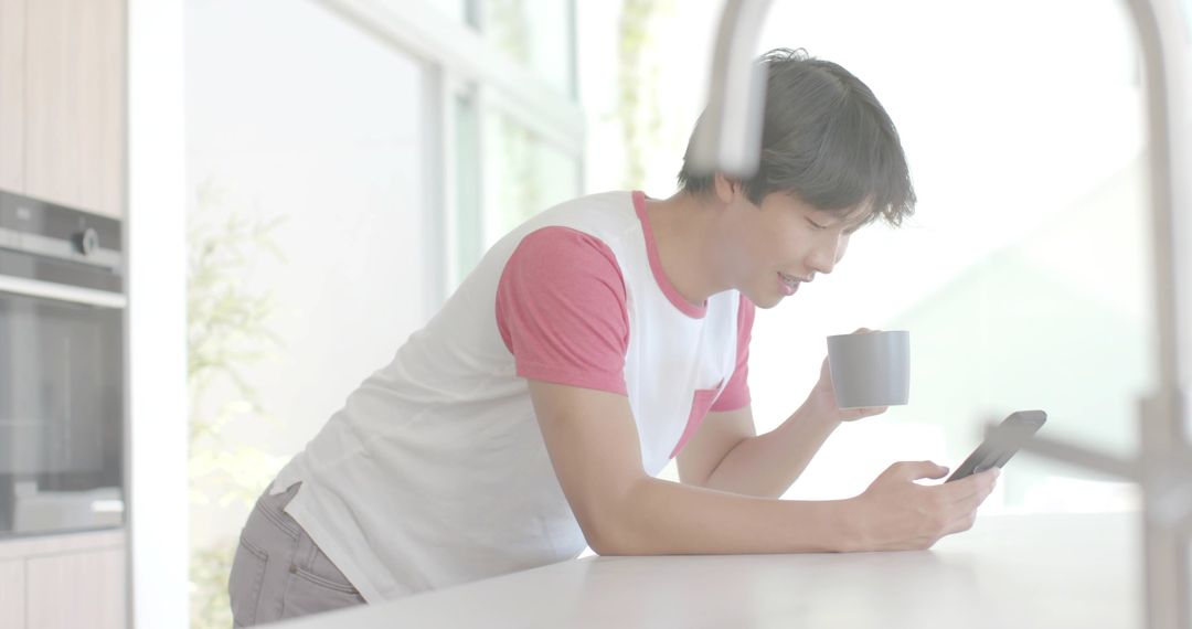 Leaning Asian man holding mug and checking phone during casual morning in bright modern kitchen