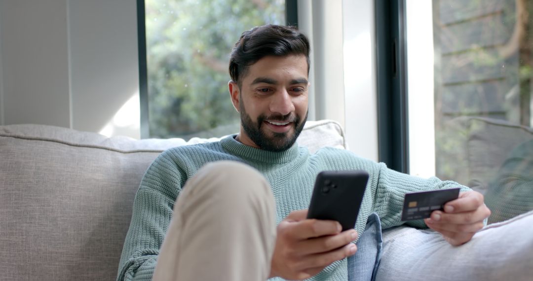 Smiling Man Shopping Online with Smartphone and Credit Card on Couch