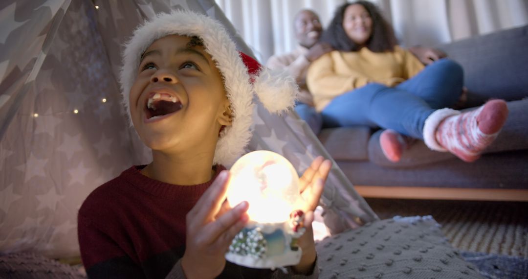 Child Holding Christmas Snow Globe While Parents Watch In Background - Free Images, Stock Photos and Pictures on Pikwizard.com