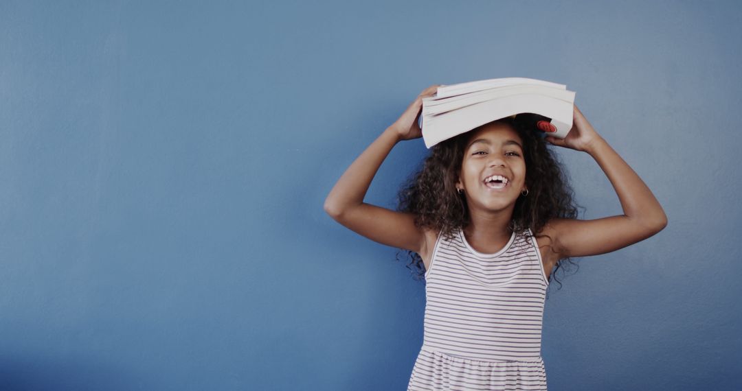 Happy Girl Balancing Books on Head in Front of Blue Wall - Free Images, Stock Photos and Pictures on Pikwizard.com