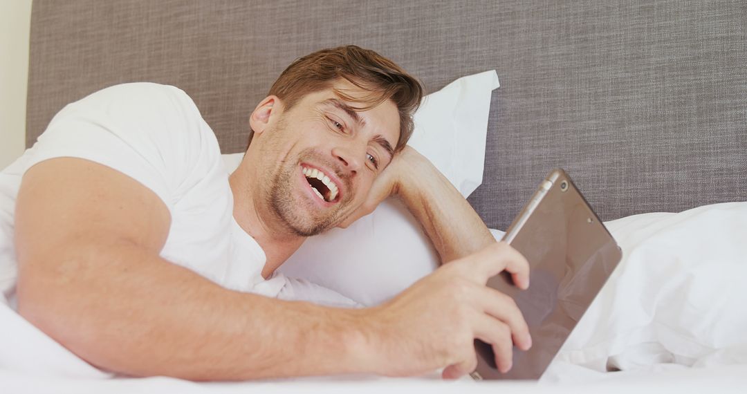 Young Man Relaxing in Bed Laughing While Using Tablet - Free Images, Stock Photos and Pictures on Pikwizard.com