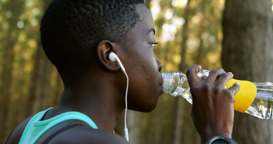 Tired african american sportswoman with headphones, drinking water in forest on sunny day - Free Images, Stock Photos and Pictures on Pikwizard.com