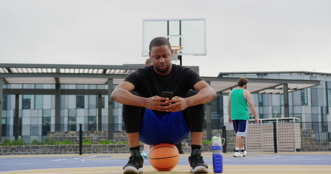 Young man taking a break from playing basketball while using smartphone on outdoor court - Free Images, Stock Photos and Pictures on Pikwizard.com