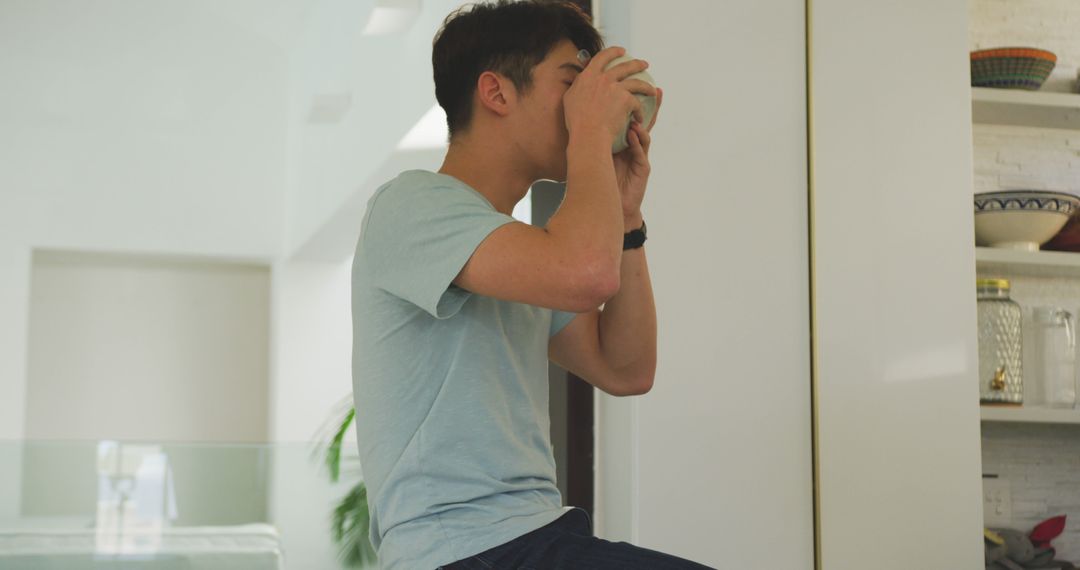 Young Man Drinking Coffee Sititing on Counter in Bright Modern Kitchen - Free Images, Stock Photos and Pictures on Pikwizard.com