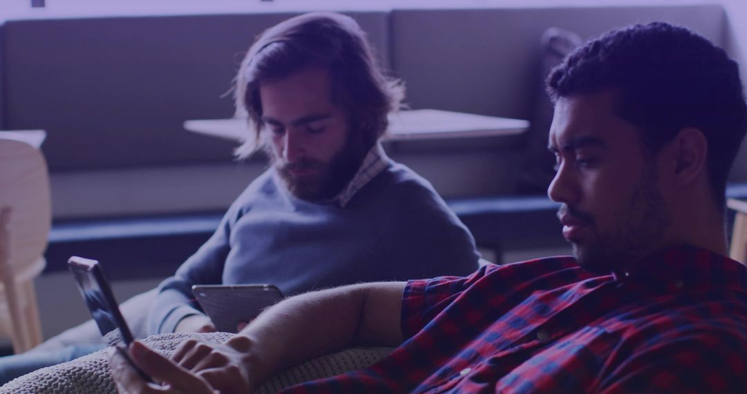 Two men lounging on couch using smartphone and tablet, casual digital downtime and coworking vibe