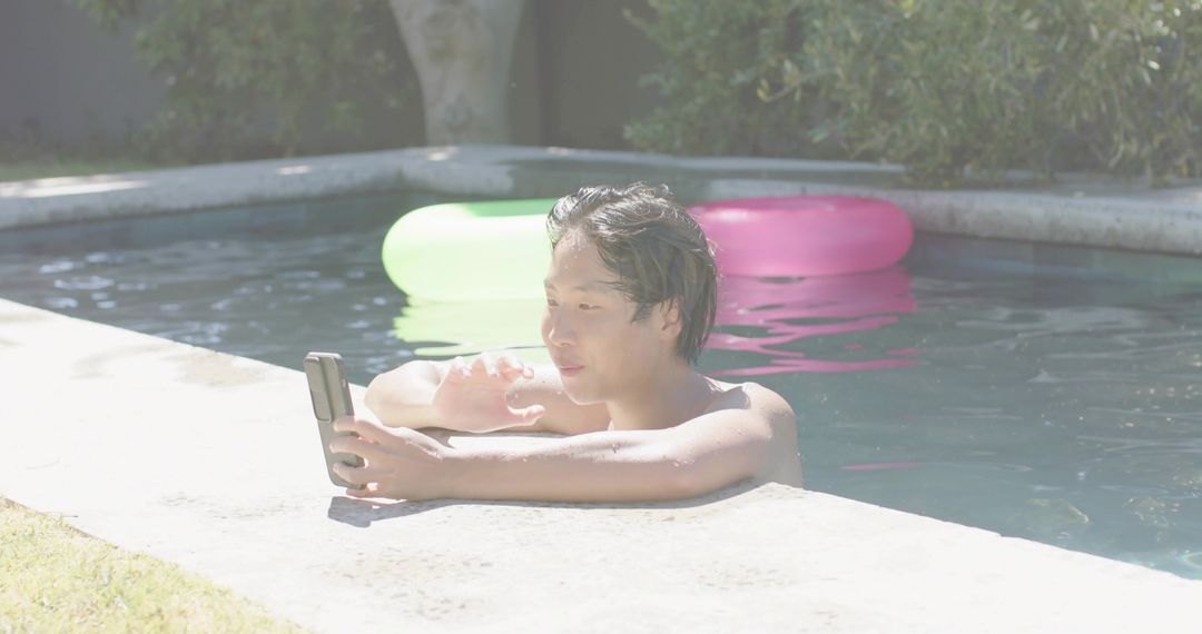 Shirtless Asian man relaxing at pool edge scrolling smartphone with neon float rings