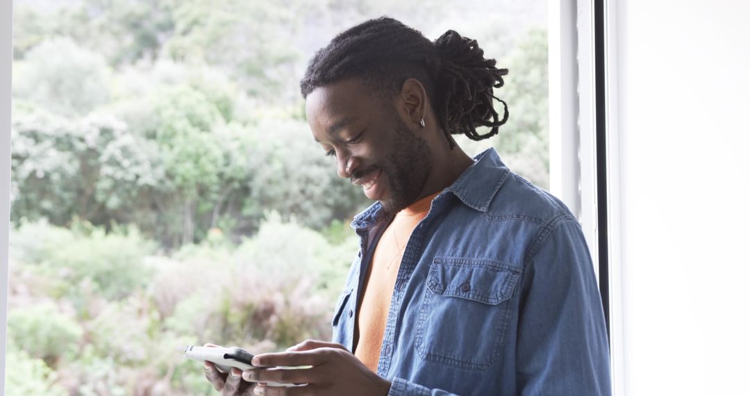 African American man standing by window smiling while using smartphone in bright interior