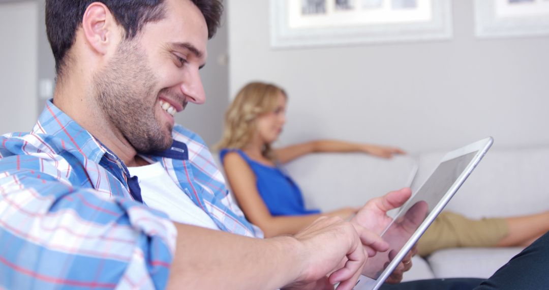 Smiling Man Using Tablet While Woman Relaxing on Sofa in Background - Free Images, Stock Photos and Pictures on Pikwizard.com