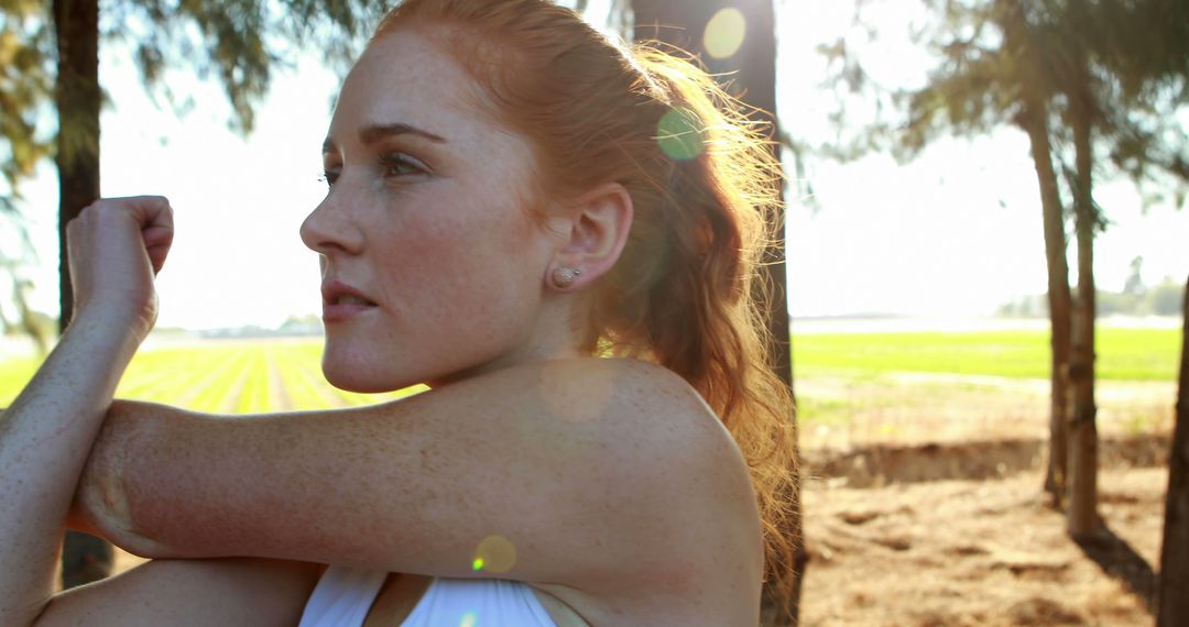 Focused redheaded woman stretching arm outdoors in sunny park - Free Images, Stock Photos and Pictures on Pikwizard.com