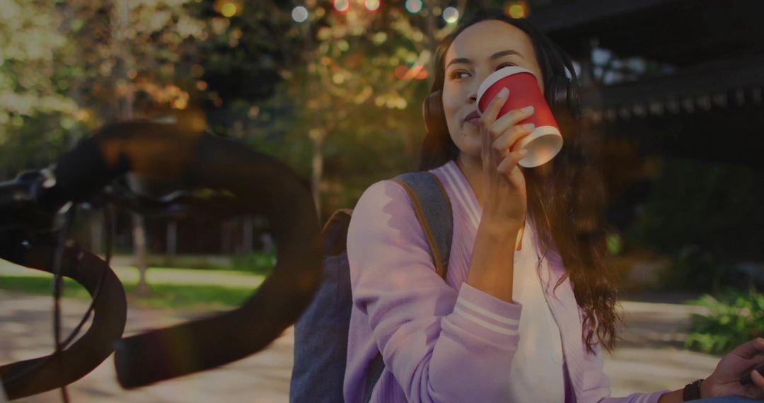 Student sipping coffee while listening to music on sunny campus with bicycle and backpack