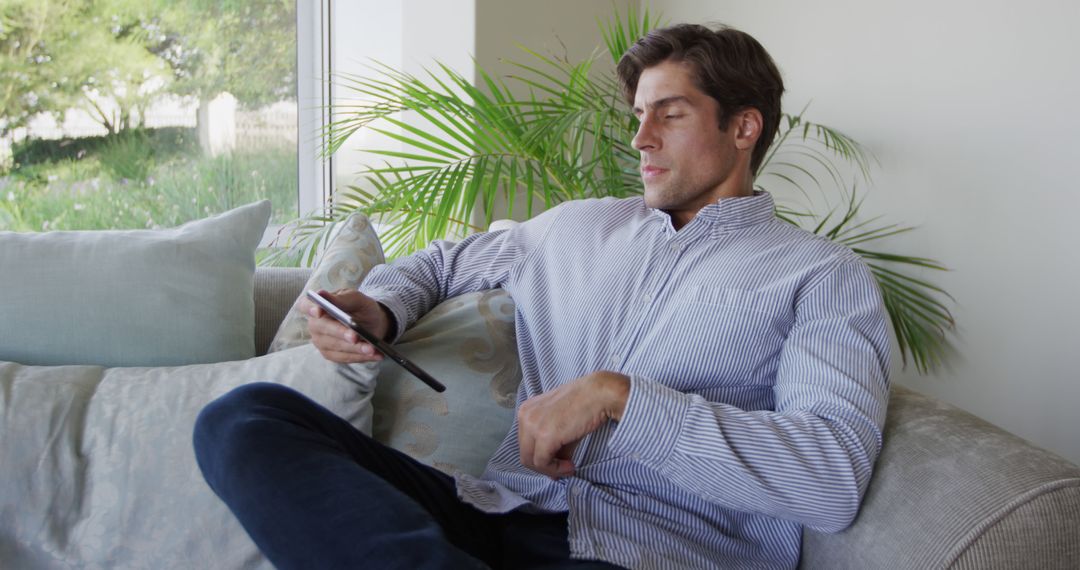 Man relaxing on couch browsing smartphone by sunny window at home - Free Images, Stock Photos and Pictures on Pikwizard.com