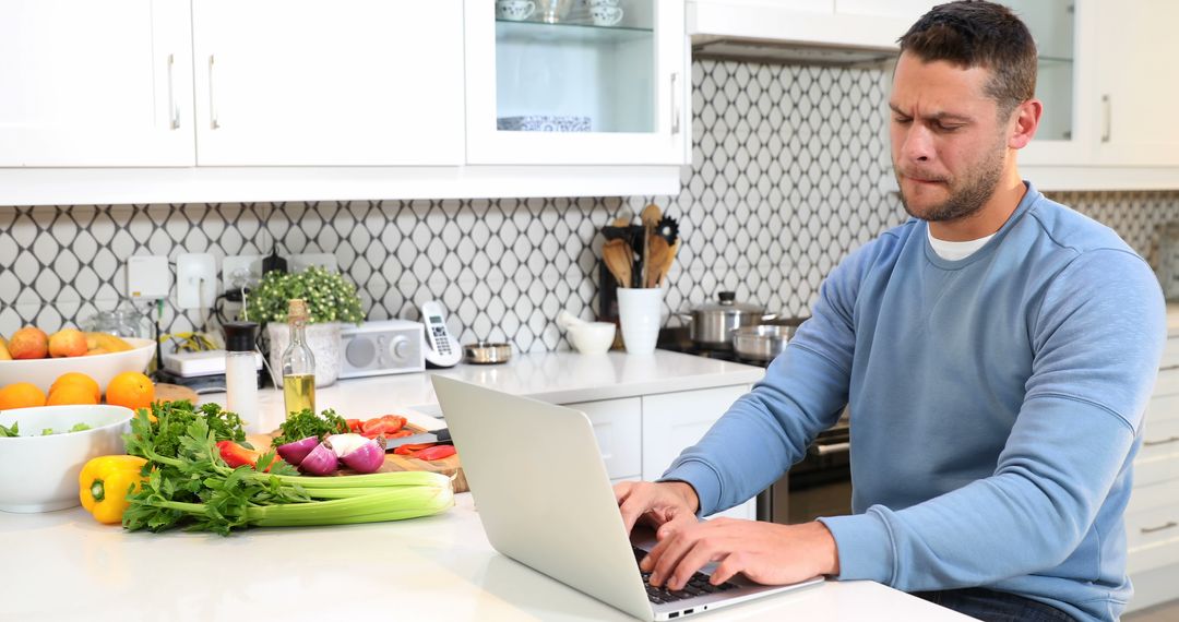 Man working on laptop in kitchen while preparing vegetables - Free Images, Stock Photos and Pictures on Pikwizard.com