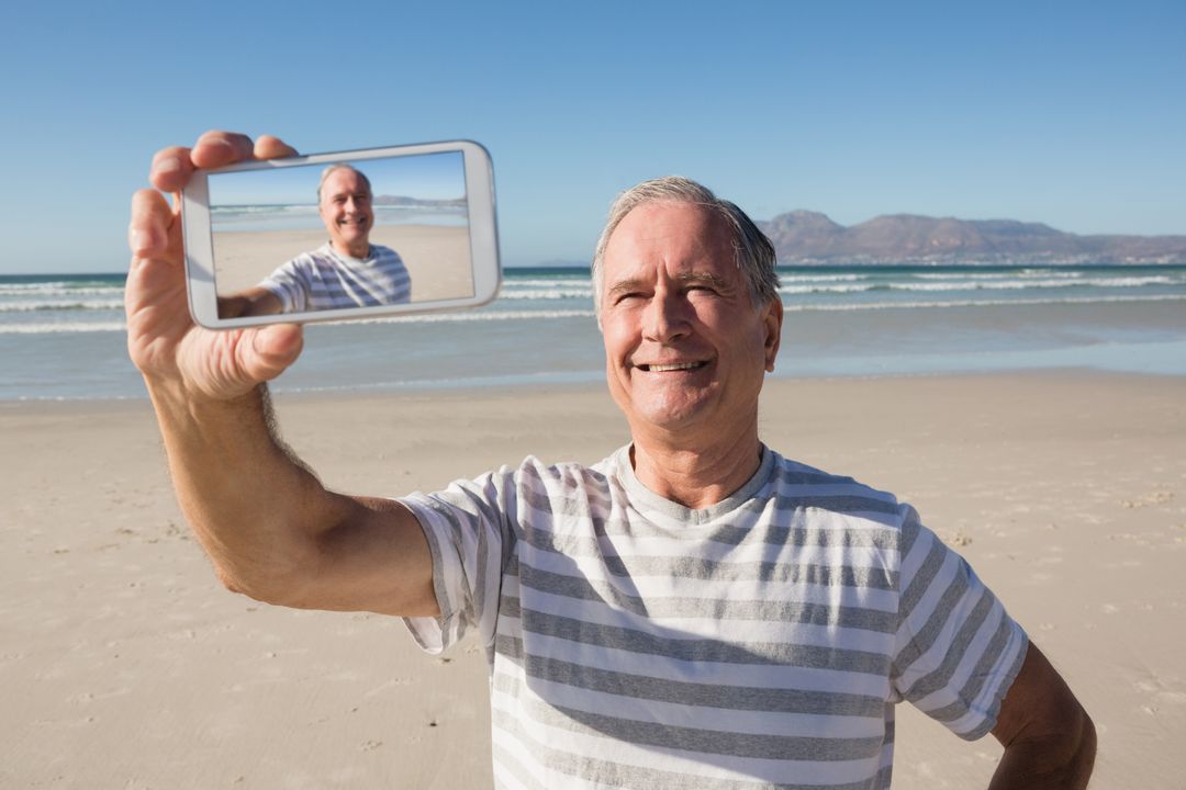 Smiling senior man holding smart phone while standing against sea - Free Images, Stock Photos and Pictures on Pikwizard.com