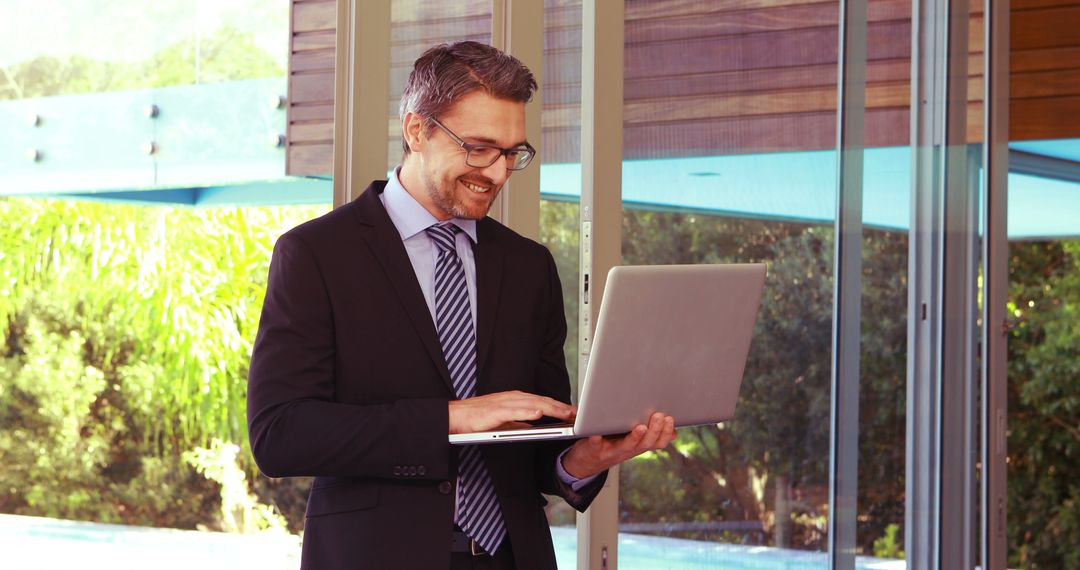 Businessman Smiling While Working on Laptop Near Large Window - Free Images, Stock Photos and Pictures on Pikwizard.com