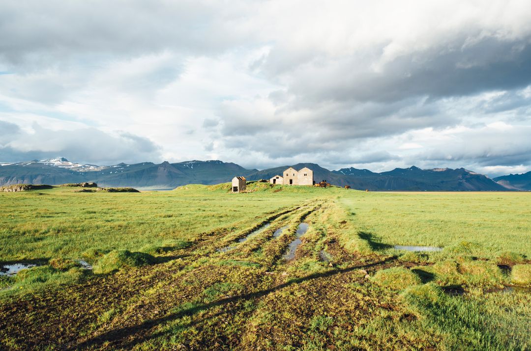 Abandoned farmhouse standing amid green marshland with muddy tracks to mountains