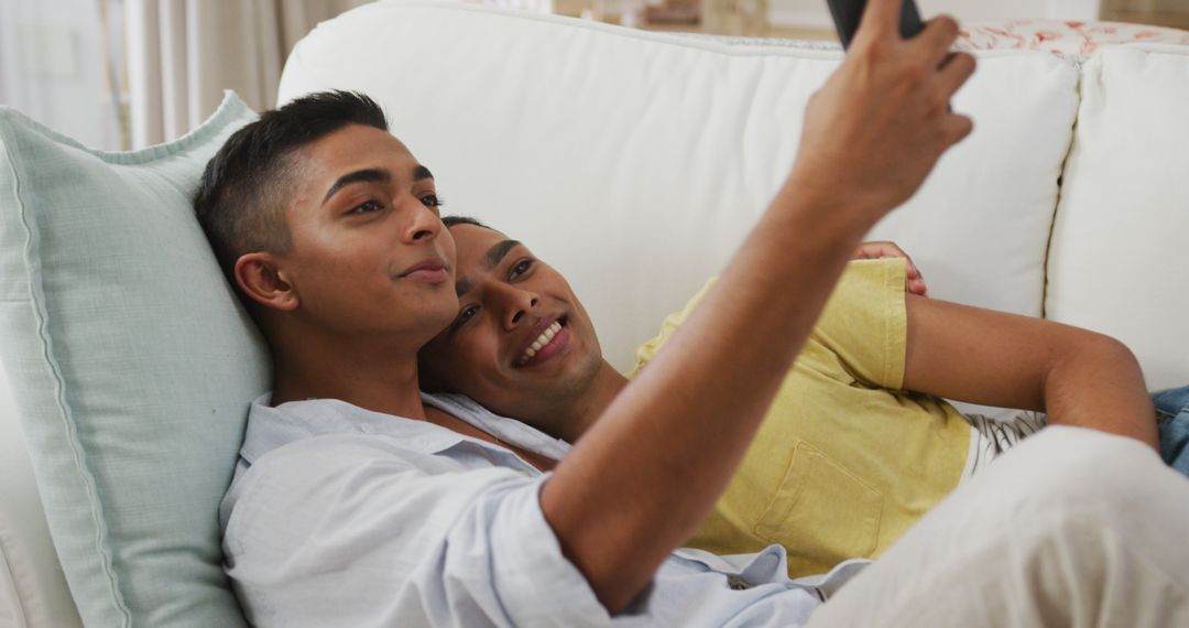 Smiling biracial gay male couple hugging lying on sofa taking selfie - Free Images, Stock Photos and Pictures on Pikwizard.com