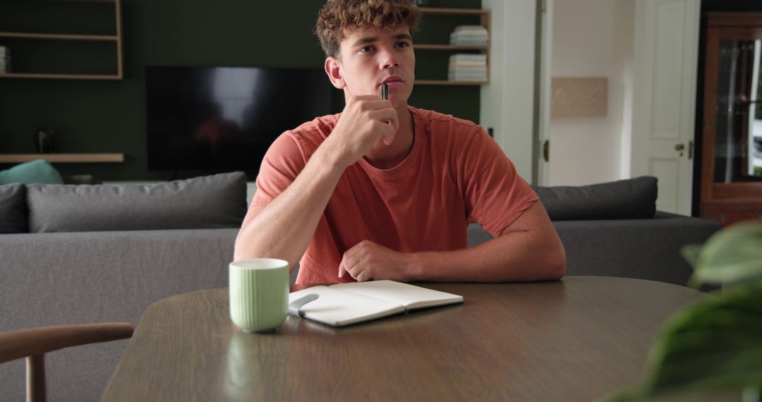 Young man sitting at table thinking with notebook and mug, home study and planning